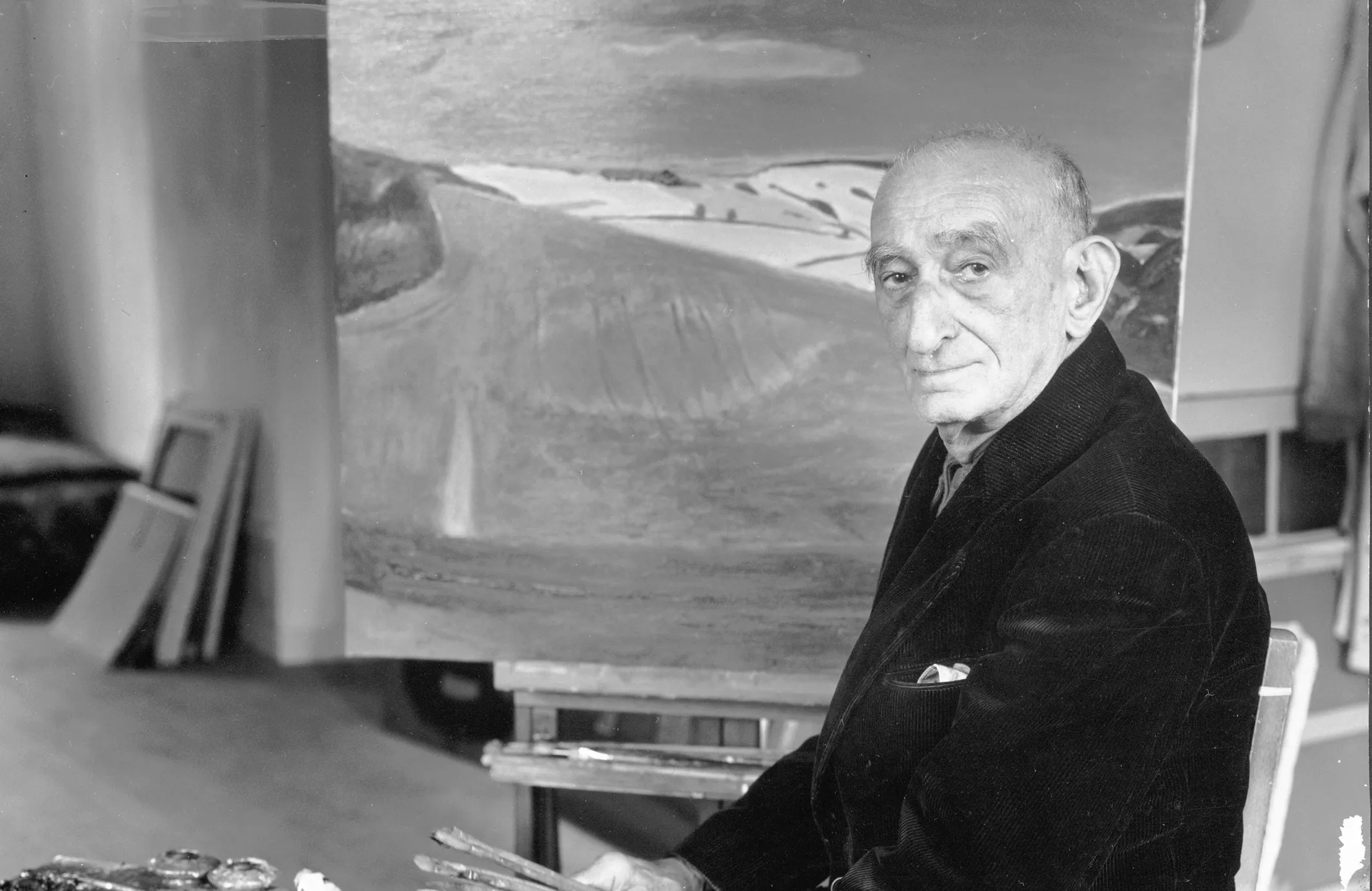 Henri Hayden assis sur une chaise avec un tableau inachevé derrière lui.